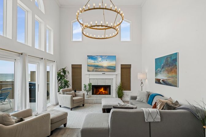 Sunlit coastal living room with vaulted ceilings and double-ring chandelier, fireplace with TV, gray sectional and armchairs, large windows and sliding doors opening to an ocean-view balcony.