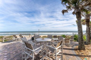 Oceanfront patio with round white table and cushioned chairs, lounge chairs facing the sandy beach, palm trees and gentle waves under a bright blue sky