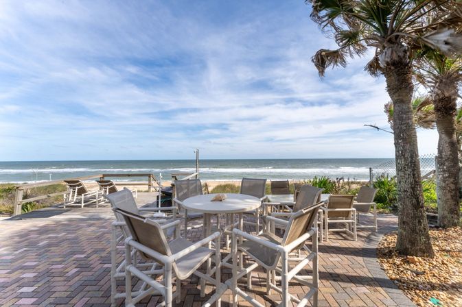 Oceanfront patio with round white table and cushioned chairs, lounge chairs facing the sandy beach, palm trees and gentle waves under a bright blue sky