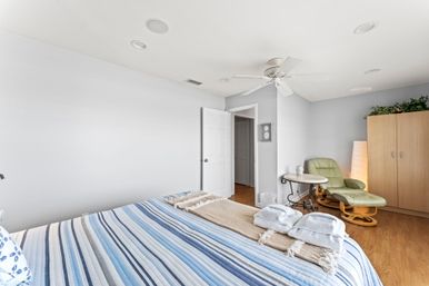 Bright, airy bedroom with blue-striped bed and folded towels, ceiling fan, green recliner and ottoman, wooden wardrobe and hardwood floors.