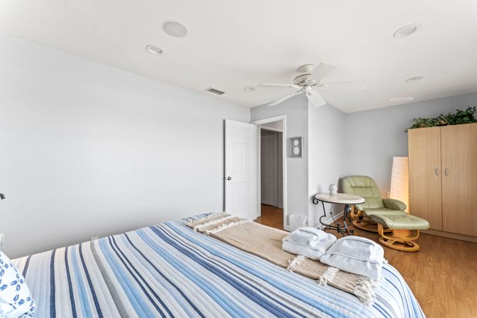 Bright, airy bedroom with blue-striped bed and folded towels, ceiling fan, green recliner and ottoman, wooden wardrobe and hardwood floors.