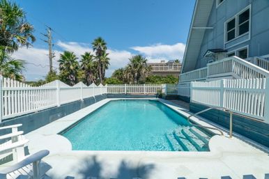 Sunny coastal vacation rental with a turquoise rectangular swimming pool, white picket fence, palm trees, and an A-frame house with deck and steps