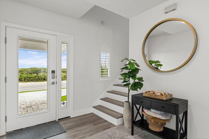 Bright modern coastal-inspired foyer with glass front door and sidelights, light wood floors, short staircase, black console table with wicker baskets and seashell tray, tall potted plant and round gold mirror.