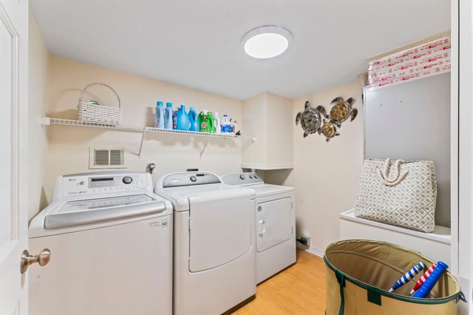 Bright home laundry room with white washer and dryer, wire shelf stocked with detergent and a wicker basket, decorative turtle wall art, patterned tote on a bench, and a round hamper on a wood floor.