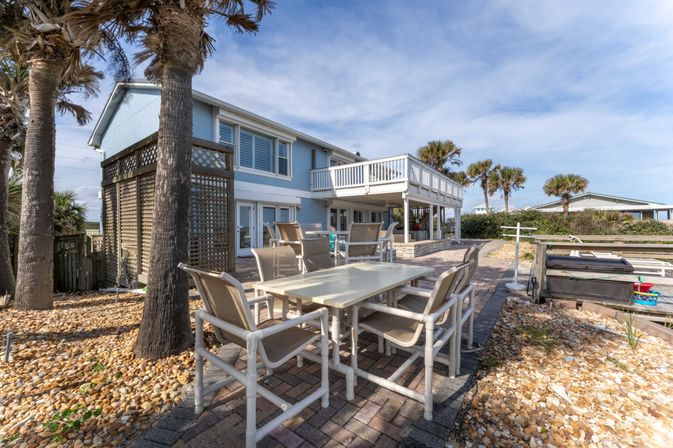 Beachfront two-story blue coastal home with palm trees, upper deck, patio dining set and ocean-view yard