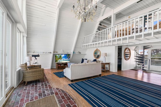 Sunlit coastal A-frame beach house living room with white vaulted wood ceiling, large ocean-view windows, wicker chair, cream sofa, blue striped rugs, chandelier, TV and spiral staircase up to a loft