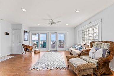 Bright beachfront living room in a coastal home with wicker armchairs and ottoman, striped cushions, a sofa with nautical pillows, hardwood floors and textured rug, ceiling fan and glass-block window, sliding glass doors opening to a sun deck with turquoise Adirondack chairs and ocean view.