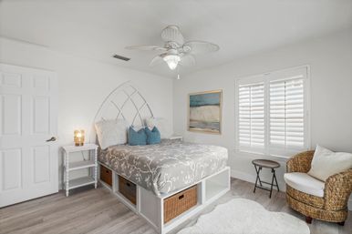 Airy coastal-style bedroom with a raised white storage platform bed and wicker baskets, gray floral duvet, blue accent pillows, arched headboard, ceiling fan, white plantation shutters, wicker armchair and light wood floors.