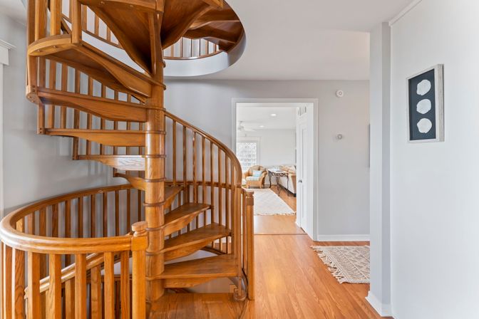 Warm wooden spiral staircase in a bright home foyer with hardwood floors and an open doorway to a sunlit living room