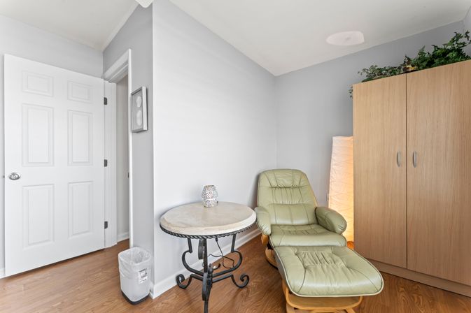 Inviting home reading nook in a quiet corner: light-green leather recliner with ottoman, round marble-top side table on wrought-iron base, tall paper floor lamp, wooden wardrobe, hardwood floor and soft gray walls.