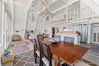 Sunlit coastal A-frame beach house interior with open-plan dining and living area, dark wooden dining table, white shiplap walls, loft balcony, chandelier, wicker seating and floor-to-ceiling glass doors.