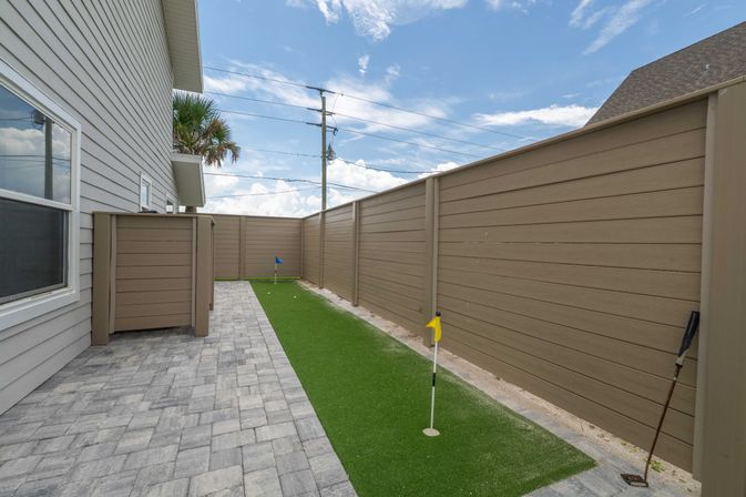 Narrow residential backyard with artificial turf putting green and two flag holes, paver patio, tall brown privacy fence, golf club leaning against fence, gray house siding and palm tree under a blue sky.