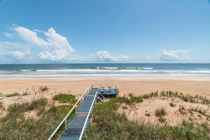 Wooden boardwalk and small viewing deck over grassy dunes leading to a wide sandy beach and calm ocean under a bright blue sky with puffy clouds