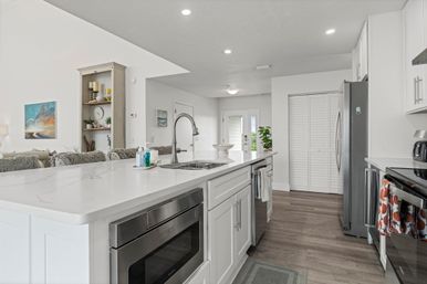 Bright, airy open-plan kitchen with large white quartz island, undermount sink and gooseneck faucet, stainless steel appliances and microwave drawer, white cabinets, wood-look floors, recessed lighting, and adjacent living area with shelving and coastal artwork