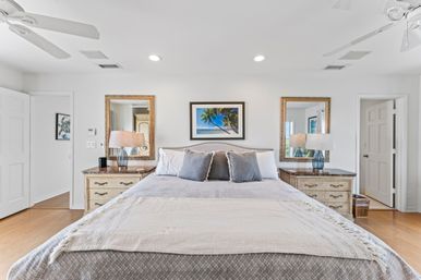 Bright coastal master bedroom with king bed, gray and white bedding, two wooden nightstands with blue glass lamps, framed tropical beach print above headboard, matching gold mirrors, ceiling fans and hardwood floor.