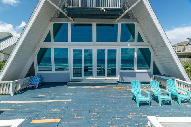 Oceanfront A-frame beach house deck with weathered blue planks, a wall of glass reflecting waves and sky, and three cheerful turquoise Adirondack chairs on a sunny day.