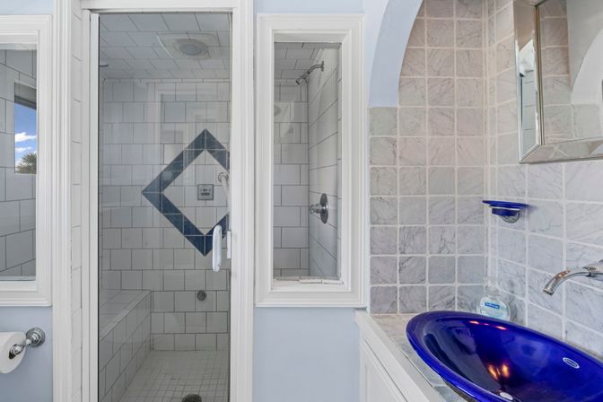 Glass-enclosed white-tile shower with blue diamond tile accent and bench, adjacent to marble mosaic wall and glossy cobalt-blue vessel sink with chrome faucet.