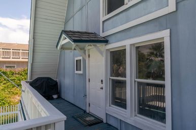 Beachside balcony entrance to a light-blue cottage with a white door, small awning, covered grill, and palm-reflecting windows.