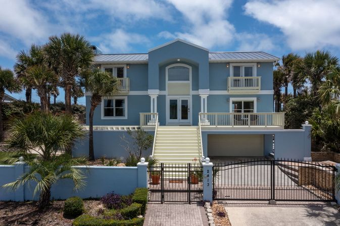 Bright blue two-story elevated coastal beach house with palm trees, wide central staircase to double doors, second-floor balconies, garage underneath and a gated driveway