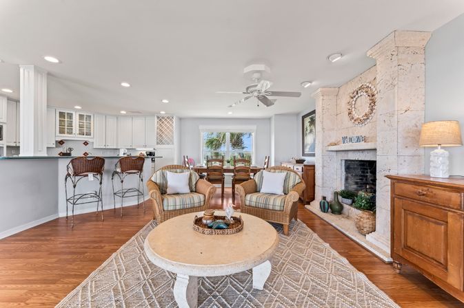 Sunny coastal living room with wicker armchairs and striped cushions, round stone coffee table on a patterned rug, white kitchen with bar stools, dining table by a window framing palm trees and ocean view, stone fireplace, ceiling fan and hardwood floors.