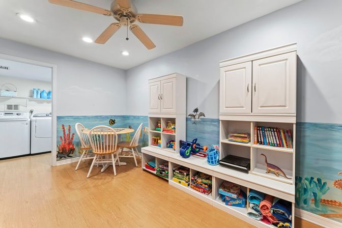 Bright children's playroom with ocean-themed wall mural, round wooden kid's table and chairs, white cubby cabinets stocked with toys and books, ceiling fan, and adjacent laundry area with washer and dryer.