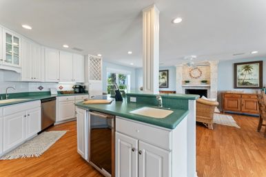 Sunny open-concept coastal kitchen and living area with white cabinets, green counters and island sink, stainless dishwasher, built-in wine rack, hardwood floors, and a stone fireplace with palm artwork.