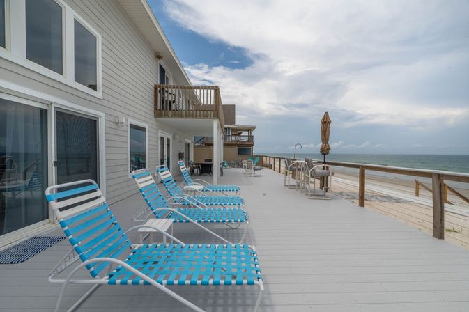 Beachfront deck of a coastal vacation home with a row of blue woven lounge chairs, bar stools and a closed umbrella along a wooden railing, overlooking a sandy beach and calm ocean under a partly cloudy sky.