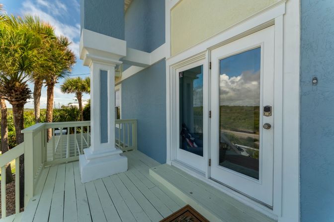 Sunlit coastal porch of a blue beach house with white French doors, pale wood deck, columned railing and palm trees reflected in the glass.