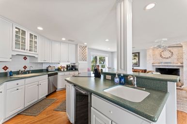 Bright, open-plan coastal-style kitchen with white cabinets, green countertops, center island with sink, stainless dishwasher, built-in wine rack, hardwood floors and palm-tree view to a living room with stone fireplace.