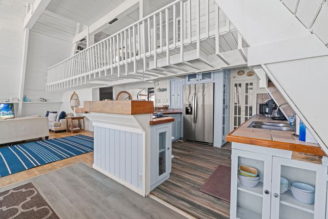 Bright coastal loft beach cottage interior with open-plan kitchen and living area, white A-frame beams, blue cabinetry, wood countertops, stainless fridge, striped navy rug and mezzanine balcony