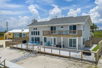 Beachfront two-story coastal house with upper balcony and furnished sun deck, turquoise lounge chairs on sandy beach under bright blue sky with puffy clouds