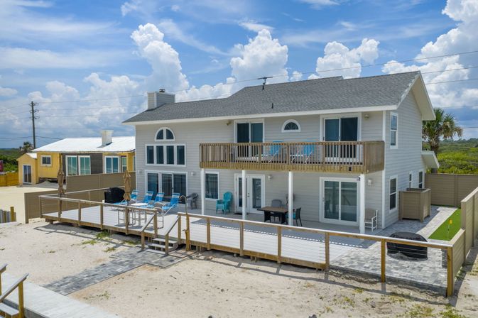 Beachfront two-story coastal house with upper balcony and furnished sun deck, turquoise lounge chairs on sandy beach under bright blue sky with puffy clouds