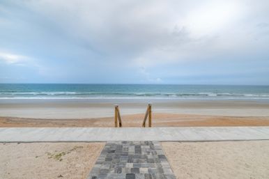 Oceanfront beach with paver walkway and wooden steps leading down to a wide sandy shoreline, gentle waves and a pale cloudy sky.
