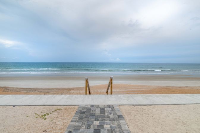 Oceanfront beach with paver walkway and wooden steps leading down to a wide sandy shoreline, gentle waves and a pale cloudy sky.