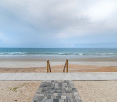 Oceanfront beach with paver walkway and wooden steps leading down to a wide sandy shoreline, gentle waves and a pale cloudy sky.
