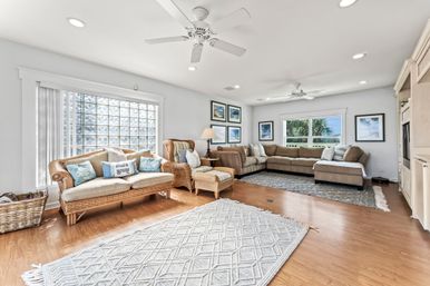 Bright coastal living room with rattan sofa and armchair, large beige sectional, hardwood floors, white ceiling fans, sea-themed pillows and artwork, and a palm-tree view through the windows.