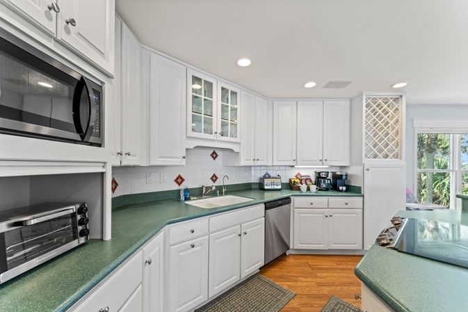 Bright coastal-style kitchen with white cabinets, green countertops, stainless microwave and dishwasher, wine rack and coffee/toaster station, plus a sunlit window showing palm trees.