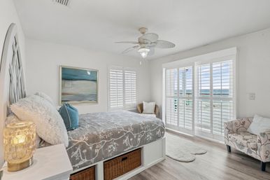 Airy coastal bedroom with ocean view through white plantation shutters, queen bed with gray floral duvet, wicker storage baskets, tropical ceiling fan and patterned accent chair