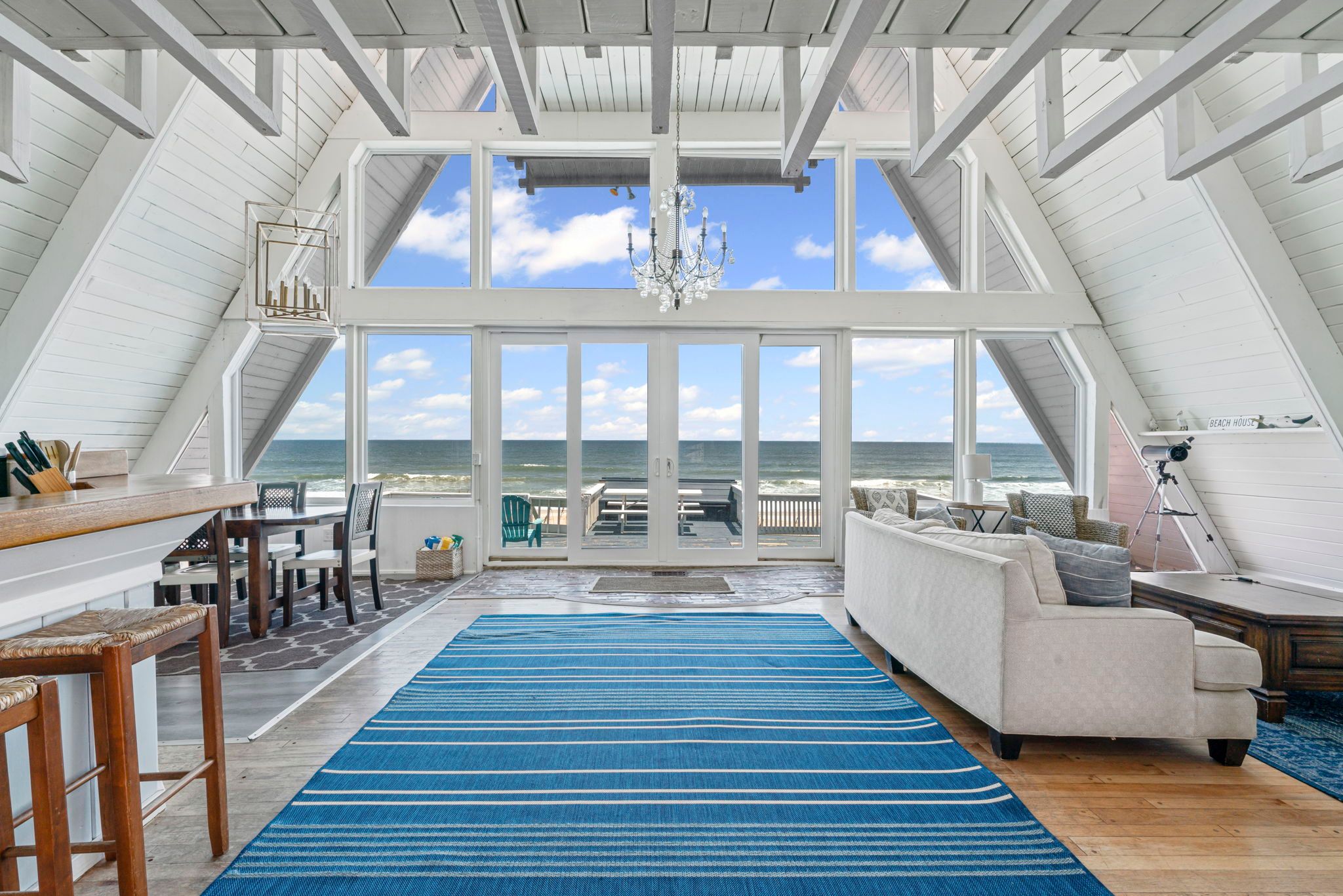 Bright A-frame beachfront living room with vaulted white beams, blue striped rug, chandelier, sofa and dining area; glass doors open to a wooden deck overlooking the ocean.