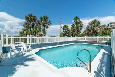 Sunlit outdoor swimming pool with clear blue water, white picket fence, palm trees in the background, metal handrail and Adirondack chairs on the deck under a bright blue sky.
