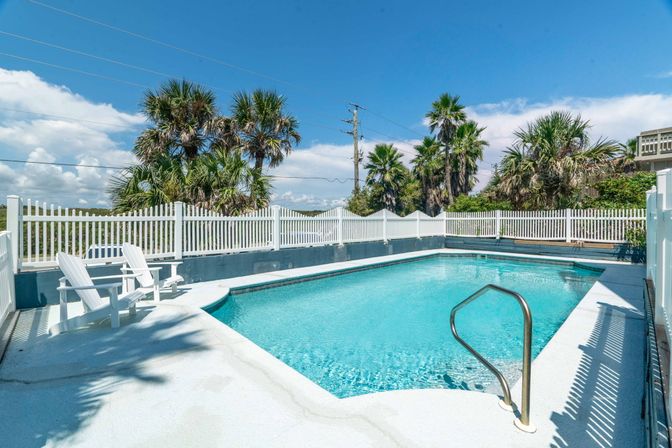 Sunlit outdoor swimming pool with clear blue water, white picket fence, palm trees in the background, metal handrail and Adirondack chairs on the deck under a bright blue sky.