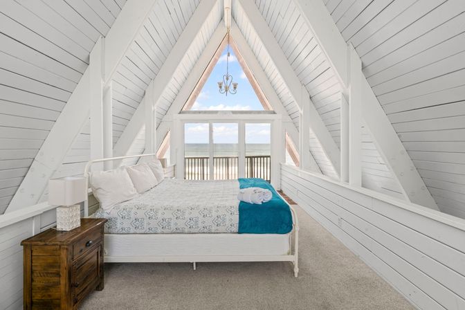 Bright white A-frame loft bedroom with shiplap walls, bed with teal throw and rolled towels, and balcony overlooking the beach and ocean