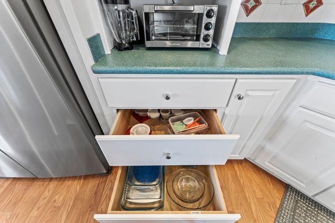 Open white kitchen drawers with organized glass mixing bowls and plastic food storage containers under a green laminate countertop with toaster oven and blender.