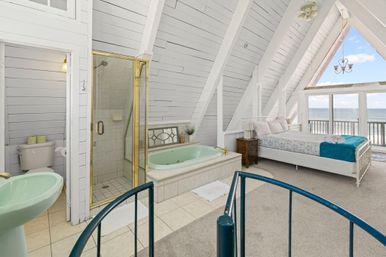 Cozy beachfront A-frame loft bedroom with vaulted white shiplap ceiling, large bed, en-suite spa bath with mint soaking tub and glass shower, spiral staircase and ocean-view balcony.