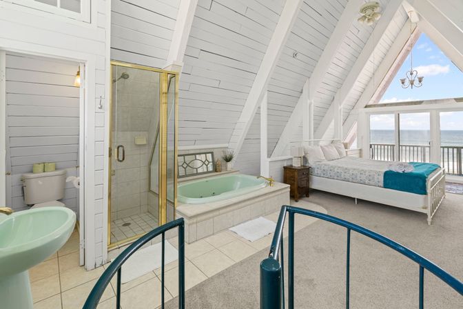 Cozy beachfront A-frame loft bedroom with vaulted white shiplap ceiling, large bed, en-suite spa bath with mint soaking tub and glass shower, spiral staircase and ocean-view balcony.