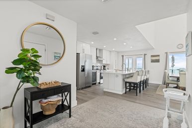 Bright open-concept coastal kitchen and dining area with a large white island and bar stools, stainless steel refrigerator and range, round wall mirror above a black console table with potted plant, hardwood floors and sliding glass doors revealing an ocean view.