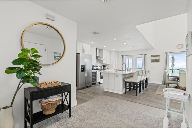 Bright open-concept coastal kitchen and dining area with a large white island and bar stools, stainless steel refrigerator and range, round wall mirror above a black console table with potted plant, hardwood floors and sliding glass doors revealing an ocean view.