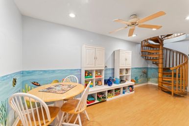 Bright basement kids' playroom with coastal underwater mural, round wooden table with white chairs, low toy-storage cabinets, ceiling fan, and a wooden spiral staircase on laminate floor