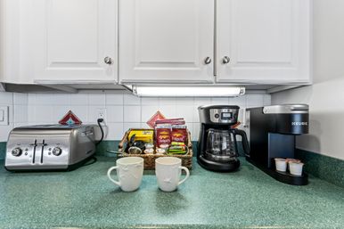 Kitchen countertop coffee station with two white mugs on a green counter, stainless toaster, drip coffee maker, single‑serve brewer, and a wicker basket of coffee and tea packets under white cabinets and a tiled backsplash.