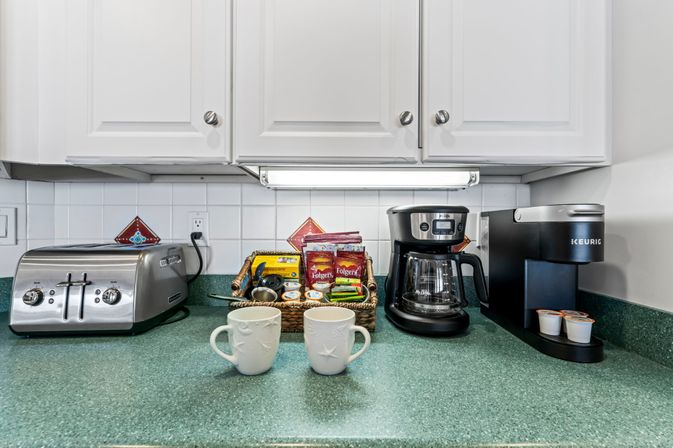 Kitchen countertop coffee station with two white mugs on a green counter, stainless toaster, drip coffee maker, single‑serve brewer, and a wicker basket of coffee and tea packets under white cabinets and a tiled backsplash.